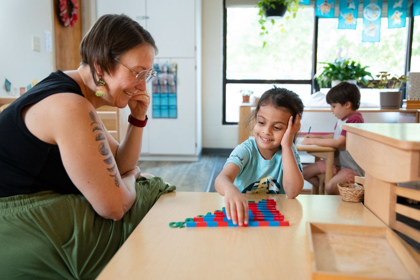 A Primary student works with Montessori math materials as her teacher observes beside her.